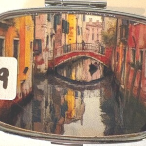 Puede incluir: Un pastillero rectangular plateado con tapa abisagrada. La caja presenta una imagen colorida y detallada de una escena de canal con un puente rojo, edificios y reflejos de agua. Una pegatina blanca con el n&uacute;mero "9" est&aacute; a la izquierda.