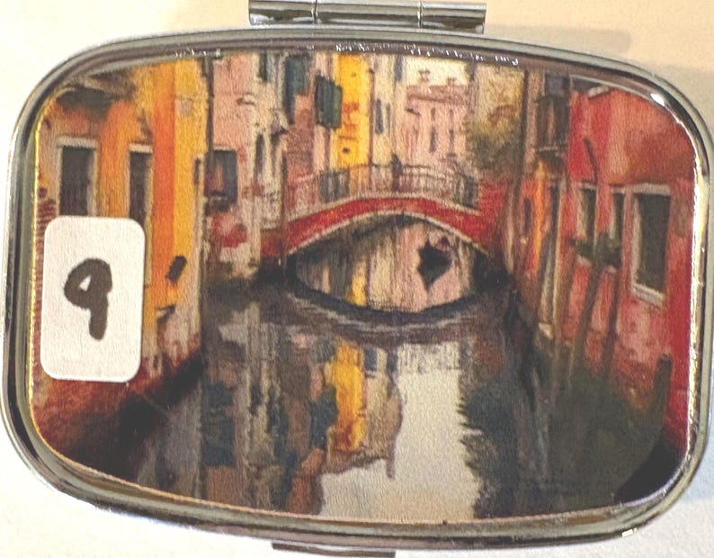 Puede incluir: Un pastillero rectangular plateado con tapa abisagrada. La caja presenta una imagen colorida y detallada de una escena de canal con un puente rojo, edificios y reflejos de agua. Una pegatina blanca con el n&uacute;mero "9" est&aacute; a la izquierda.