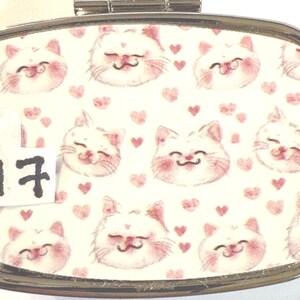 May include: A rectangular, silver-toned pill box with a hinged lid. The top features a repeating pattern of smiling white cat faces with pink cheeks and small pink hearts on a cream background. A small white tag with the number 17 is affixed to the box.