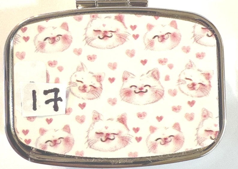 May include: A rectangular, silver-toned pill box with a hinged lid. The top features a repeating pattern of smiling white cat faces with pink cheeks and small pink hearts on a cream background. A small white tag with the number 17 is affixed to the box.