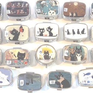 May include: A collection of small, rectangular metal pill boxes. Each box features a unique cat-themed design, including illustrations of cats with butterflies, under umbrellas, and in various artistic settings. The boxes are arranged in a grid.