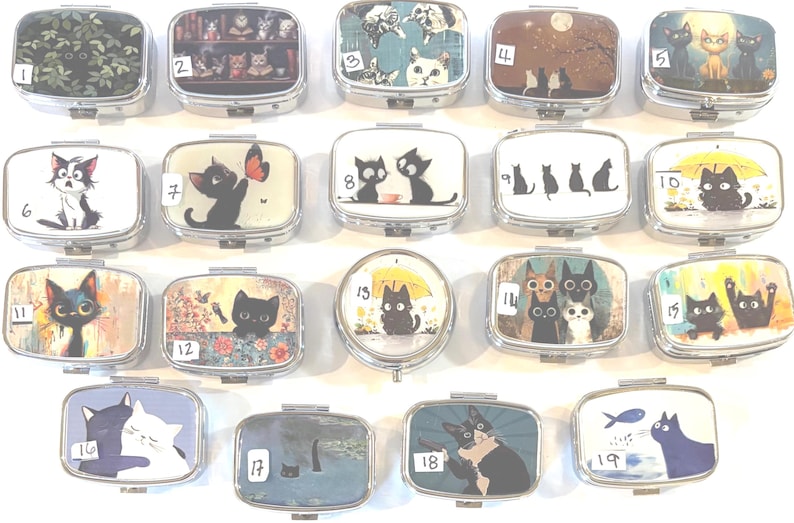 May include: A collection of small, rectangular metal pill boxes. Each box features a unique cat-themed design, including illustrations of cats with butterflies, under umbrellas, and in various artistic settings. The boxes are arranged in a grid.