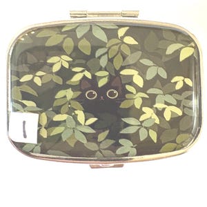 May include: A rectangular compact mirror with a silver-colored frame. The mirror's surface features a black cat peeking through green and yellow leaves. A small white sticker with the number 1 is in the lower left corner.