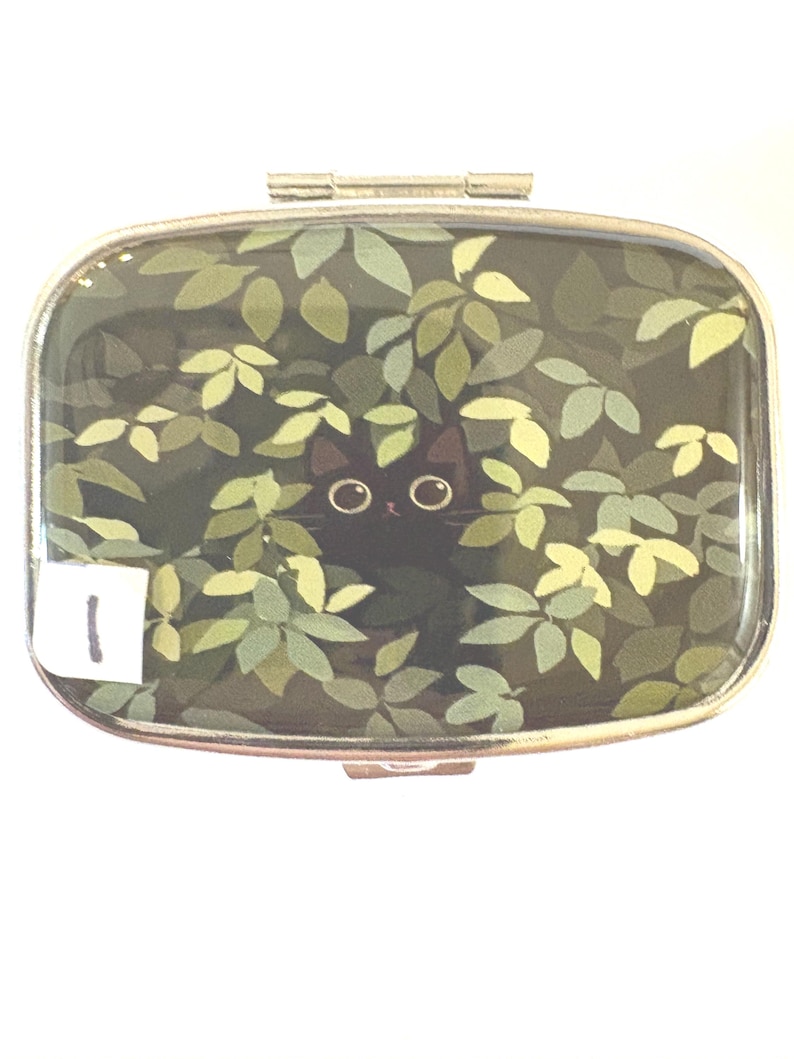 May include: A rectangular compact mirror with a silver-colored frame. The mirror's surface features a black cat peeking through green and yellow leaves. A small white sticker with the number 1 is in the lower left corner.