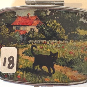 Puede incluir: Pastillero plateado con una pintura de paisaje. La pintura representa un gato negro caminando por un camino en un campo de flores, con una casa y un tejado rojo al fondo. Una pegatina con el n&uacute;mero 18 est&aacute; en la parte inferior izquierda.