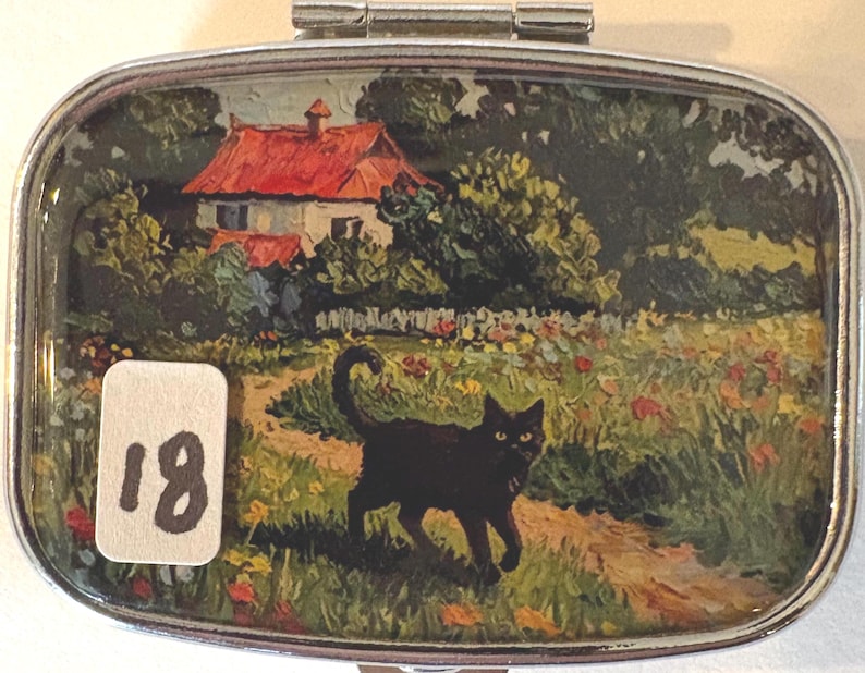 Puede incluir: Pastillero plateado con una pintura de paisaje. La pintura representa un gato negro caminando por un camino en un campo de flores, con una casa y un tejado rojo al fondo. Una pegatina con el n&uacute;mero 18 est&aacute; en la parte inferior izquierda.