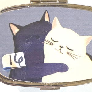 May include: A silver-toned pill box with a blue background featuring a black cat hugging a white cat. The cats are depicted with closed eyes, suggesting affection. A sticker with the number "16" is on the lower left corner.
