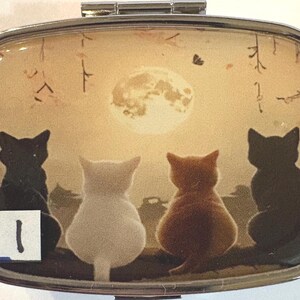 May include: A silver pill box with a rectangular shape. The lid features a serene scene of four cats silhouetted against a full moon. The cats are in various colors, including black, white, and orange.