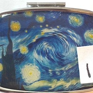 May include: A rectangular metal pill case with a hinged lid featuring a printed reproduction of a swirling blue and yellow night sky painting. A small white rectangular paper tag with the handwritten number 1 is attached to the right side.