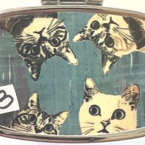 May include: A rectangular, silver-toned pill box featuring a teal background with four cat faces. The cats are in various positions, with two upside down. A small sticker with the number "3" is in the lower left corner.