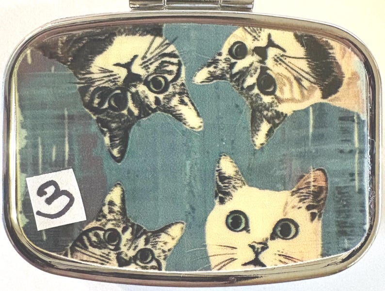 May include: A rectangular, silver-toned pill box featuring a teal background with four cat faces. The cats are in various positions, with two upside down. A small sticker with the number "3" is in the lower left corner.