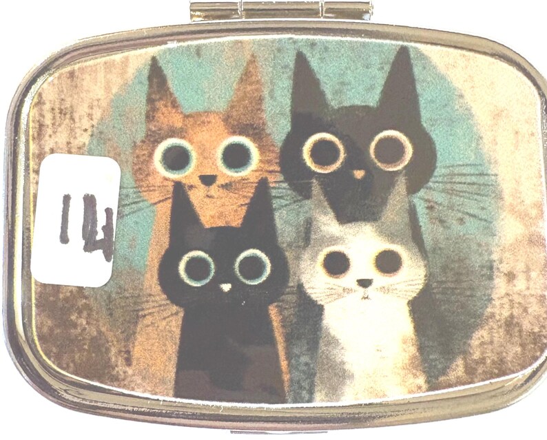 May include: A small, rectangular pill box with a hinged lid. The box features a whimsical illustration of four cats with large, round eyes in shades of orange, black, and gray against a teal background. A white sticker with the number 14 is on the left.