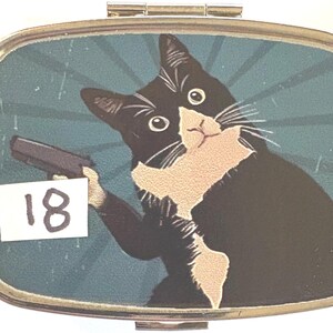May include: A rectangular pill box with a hinged lid. The box features a cartoon image of a black and white cat holding a gun against a teal background with radial lines. The number "18" is on a white sticker.