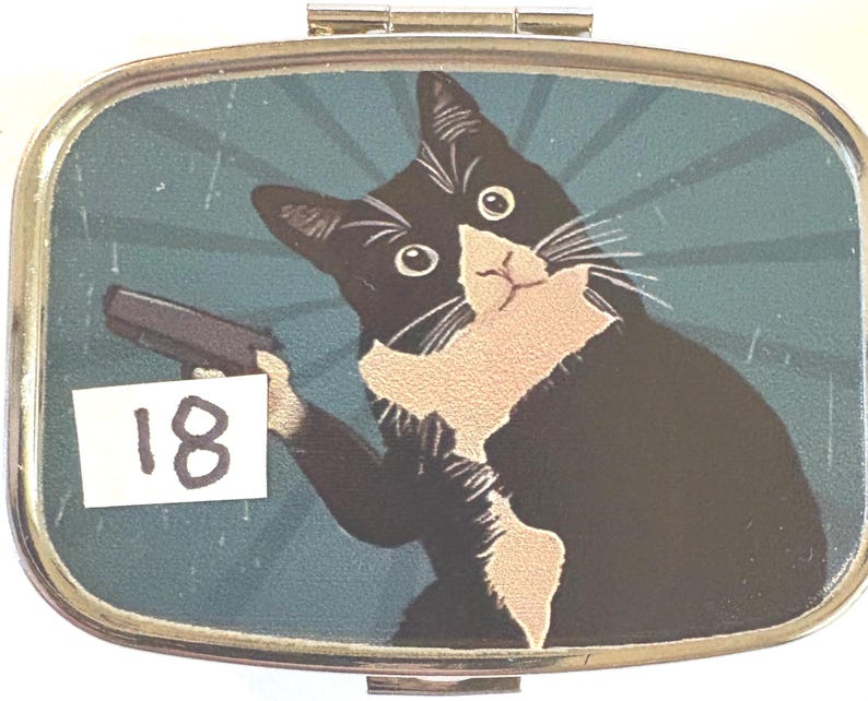 May include: A rectangular pill box with a hinged lid. The box features a cartoon image of a black and white cat holding a gun against a teal background with radial lines. The number "18" is on a white sticker.