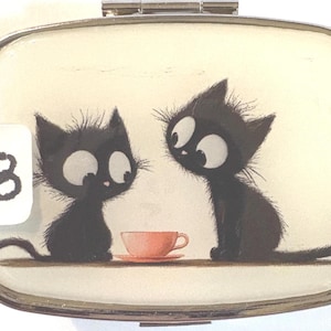 May include: A rectangular, silver-framed pill box with a whimsical illustration of two black cats with large eyes, seated near a pink teacup. A white sticker with the number 8 is in the corner. The background is a cream color.