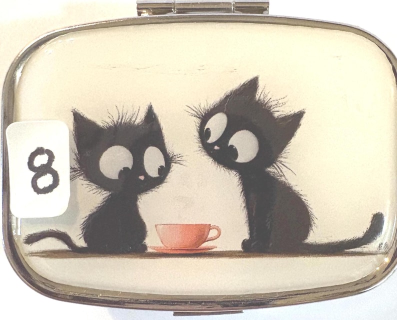 May include: A rectangular, silver-framed pill box with a whimsical illustration of two black cats with large eyes, seated near a pink teacup. A white sticker with the number 8 is in the corner. The background is a cream color.