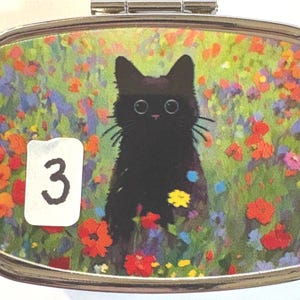 May include: A silver-toned, rectangular compact mirror with a hinged lid. The artwork features a black cat with large eyes in a field of colorful flowers. A white sticker with the number "3" is on the left.