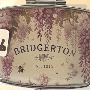Puede incluir: Un pastillero rectangular plateado con un dise&ntilde;o floral y la palabra "BRIDGERTON". El dise&ntilde;o incluye glicinas moradas, una abeja y el texto "EST. 1813". Una pegatina blanca con el n&uacute;mero "16" est&aacute; a la izquierda.