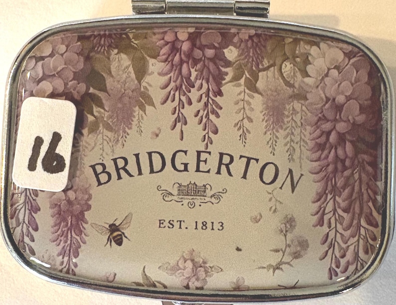Puede incluir: Un pastillero rectangular plateado con un dise&ntilde;o floral y la palabra "BRIDGERTON". El dise&ntilde;o incluye glicinas moradas, una abeja y el texto "EST. 1813". Una pegatina blanca con el n&uacute;mero "16" est&aacute; a la izquierda.
