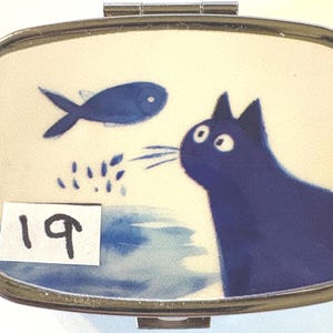 May include: A rectangular, silver-toned pill box with a hinged lid. The box features a whimsical illustration of a blue cat gazing at a blue fish. The background is a light cream color with blue accents. The number "19" is on a white sticker.