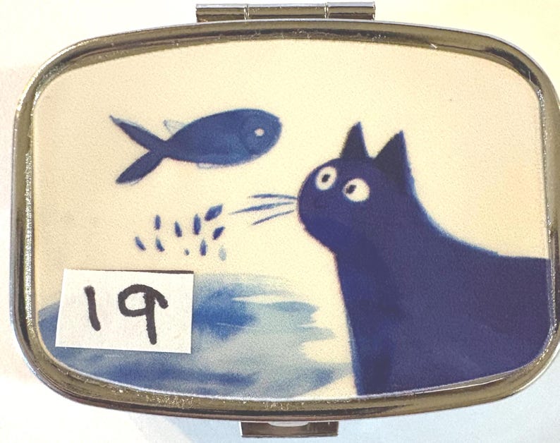 May include: A rectangular, silver-toned pill box with a hinged lid. The box features a whimsical illustration of a blue cat gazing at a blue fish. The background is a light cream color with blue accents. The number "19" is on a white sticker.