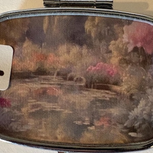 Puede incluir: Un peque&ntilde;o pastillero rectangular con un marco de metal plateado. La tapa presenta una pintura al estilo acuarela de un estanque con flores rosas y follaje verde. Una peque&ntilde;a etiqueta blanca con la letra "F" est&aacute; adjunta.