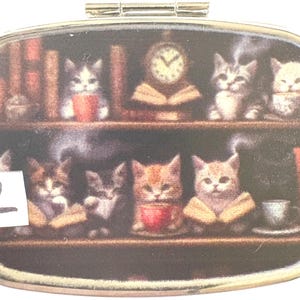 May include: An oval-shaped pill box with a decorative image of kittens on bookshelves. The kittens are depicted with books, cups, and a clock. The number "2" is visible on the left side of the box.