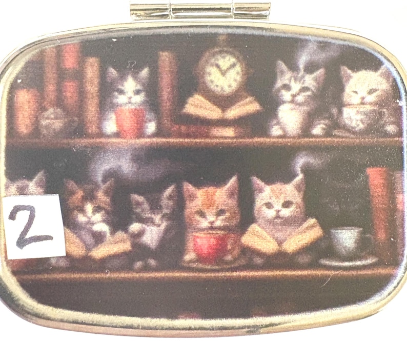 May include: An oval-shaped pill box with a decorative image of kittens on bookshelves. The kittens are depicted with books, cups, and a clock. The number "2" is visible on the left side of the box.