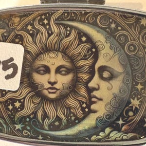 Puede incluir: Pastillero rectangular de metal con un dise&ntilde;o celestial. La obra de arte presenta un sol y una luna con caras, rodeados de estrellas y patrones arremolinados. Una pegatina con el n&uacute;mero "15" est&aacute; en la parte delantera.