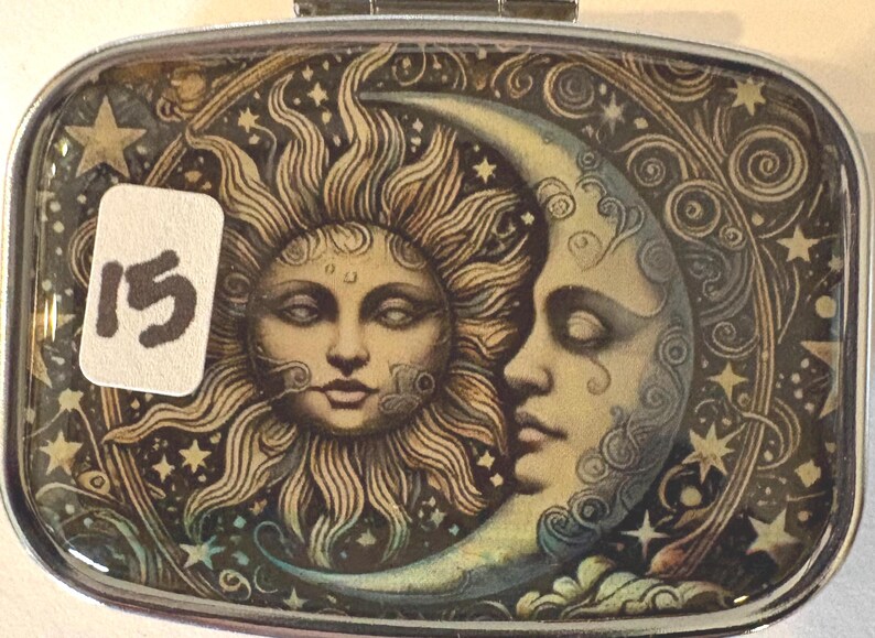 Puede incluir: Pastillero rectangular de metal con un dise&ntilde;o celestial. La obra de arte presenta un sol y una luna con caras, rodeados de estrellas y patrones arremolinados. Una pegatina con el n&uacute;mero "15" est&aacute; en la parte delantera.