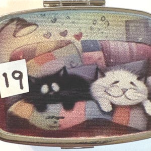 May include: A silver metal pill box with a colorful illustration of two cats lounging on a couch. A black cat and a white cat are shown. The number "19" is on a white sticker in the corner.