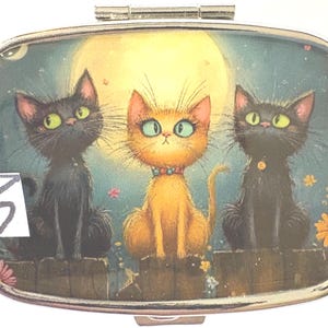 May include: A silver pill box with a whimsical illustration of three cats under a full moon. Two black cats flank an orange cat, all with large eyes. The number "5" is visible on the lower left.