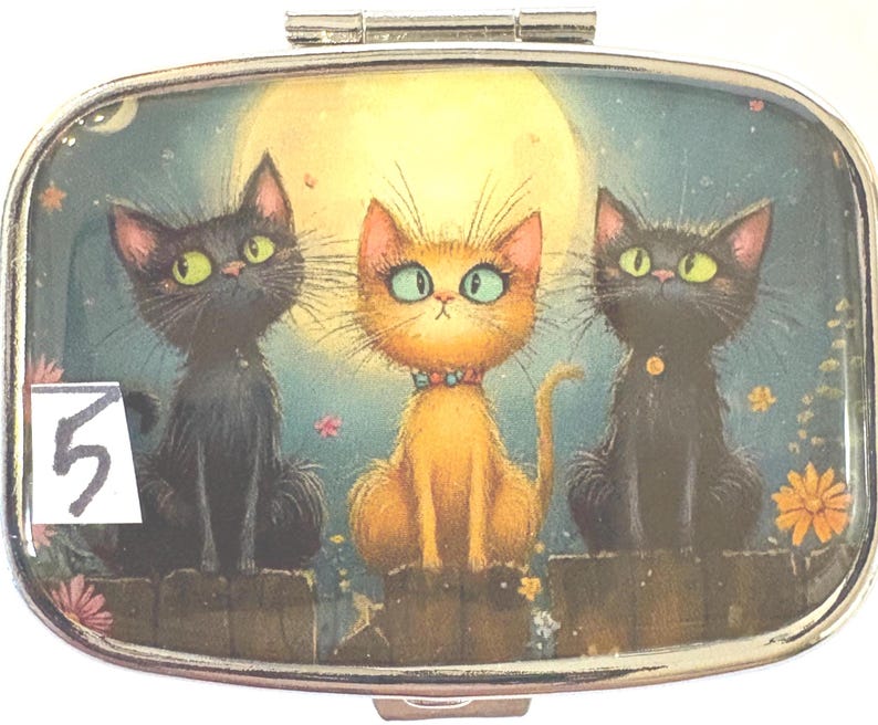 May include: A silver pill box with a whimsical illustration of three cats under a full moon. Two black cats flank an orange cat, all with large eyes. The number "5" is visible on the lower left.