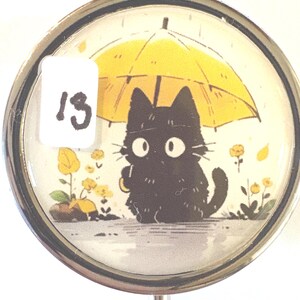 May include: A round, silver-toned pill box with a whimsical illustration of a black cat under a yellow umbrella. The cat has large, white eyes and is surrounded by yellow flowers. A white sticker with the number "13" is on the left.