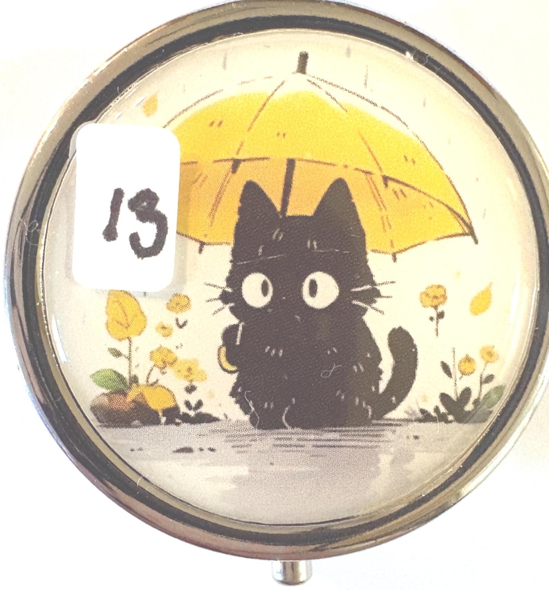 May include: A round, silver-toned pill box with a whimsical illustration of a black cat under a yellow umbrella. The cat has large, white eyes and is surrounded by yellow flowers. A white sticker with the number "13" is on the left.
