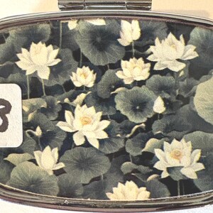 Puede incluir: Un pastillero plateado con un dise&ntilde;o floral. La caja presenta un patr&oacute;n de flores de loto blancas y hojas de nen&uacute;far verdes sobre un fondo oscuro. Una peque&ntilde;a etiqueta blanca con el n&uacute;mero 8 est&aacute; en el lado izquierdo.