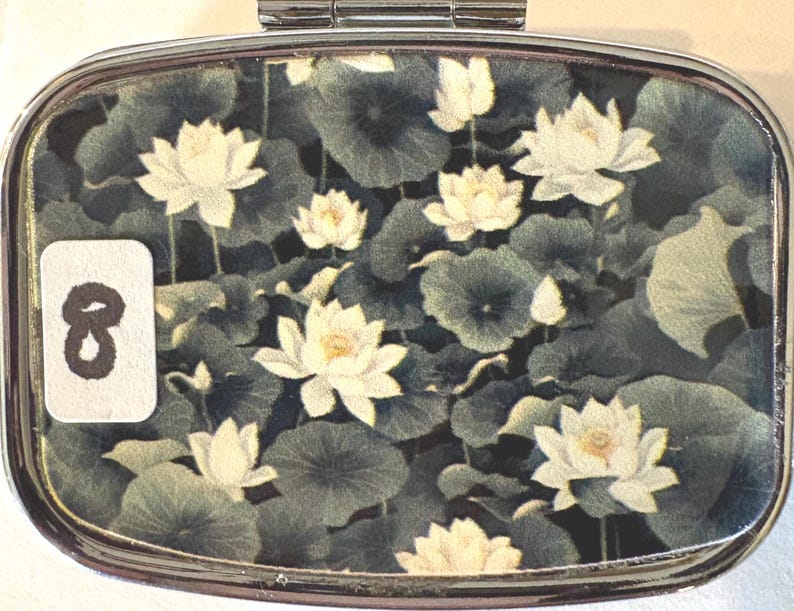 Puede incluir: Un pastillero plateado con un dise&ntilde;o floral. La caja presenta un patr&oacute;n de flores de loto blancas y hojas de nen&uacute;far verdes sobre un fondo oscuro. Una peque&ntilde;a etiqueta blanca con el n&uacute;mero 8 est&aacute; en el lado izquierdo.