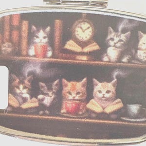 Puede incluir: Un peque&ntilde;o pastillero rectangular con un dise&ntilde;o caprichoso de gatitos en una biblioteca. La imagen muestra varios gatitos leyendo libros, bebiendo de tazas y un reloj. La paleta de colores incluye marrones c&aacute;lidos y blancos suaves.
