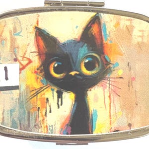 May include: A metal pill box with a colorful illustration of a black cat with large yellow eyes. The cat is set against a background of abstract paint splatters in shades of orange, yellow, and blue. The number "11" is visible on the left side.