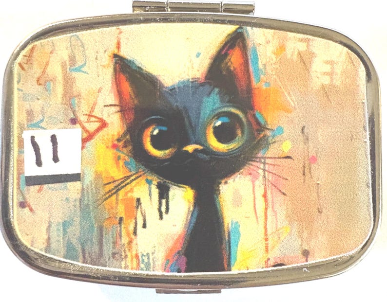 May include: A metal pill box with a colorful illustration of a black cat with large yellow eyes. The cat is set against a background of abstract paint splatters in shades of orange, yellow, and blue. The number "11" is visible on the left side.