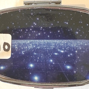 Puede incluir: Un peque&ntilde;o pastillero rectangular plateado con tapa abisagrada. El interior presenta un dise&ntilde;o azul oscuro y negro con estrellas blancas y una banda horizontal de luz. Una pegatina blanca con el n&uacute;mero "10" est&aacute; adherida a la caja.