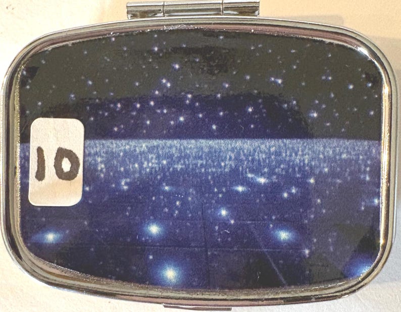 Puede incluir: Un peque&ntilde;o pastillero rectangular plateado con tapa abisagrada. El interior presenta un dise&ntilde;o azul oscuro y negro con estrellas blancas y una banda horizontal de luz. Una pegatina blanca con el n&uacute;mero "10" est&aacute; adherida a la caja.