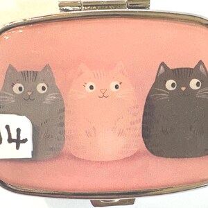 May include: A rectangular, silver-framed pill box with a pink background. The box features three cartoon cats in gray, pink, and black. A white sticker with the number 14 is on the left side of the box.