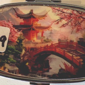 Puede incluir: Un pastillero rectangular de metal con tapa abisagrada. La tapa presenta una imagen esc&eacute;nica de una pagoda roja, un puente y flores de cerezo. Una pegatina blanca con el n&uacute;mero "19" est&aacute; en la parte delantera.