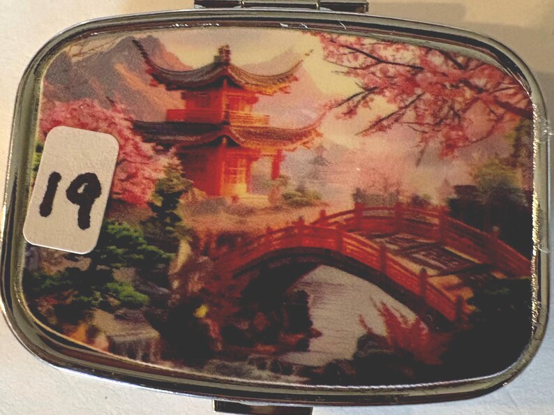 Puede incluir: Un pastillero rectangular de metal con tapa abisagrada. La tapa presenta una imagen esc&eacute;nica de una pagoda roja, un puente y flores de cerezo. Una pegatina blanca con el n&uacute;mero "19" est&aacute; en la parte delantera.