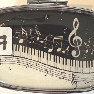 Puede incluir: Un pastillero rectangular con marco plateado con un tema musical. El dise&ntilde;o presenta un teclado de piano, notas musicales y una clave de sol sobre un fondo negro. Una pegatina blanca con el n&uacute;mero 7 est&aacute; a la izquierda.