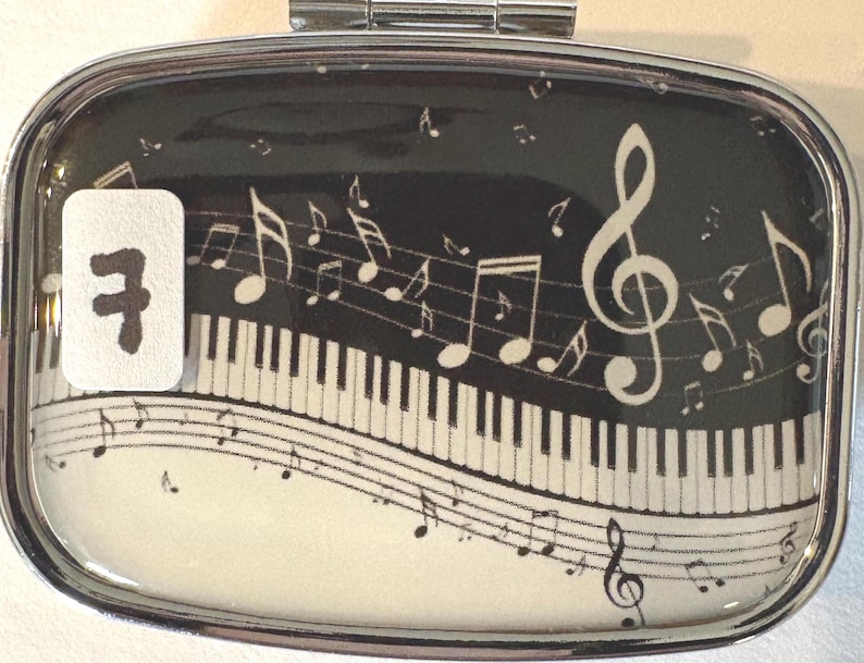Puede incluir: Un pastillero rectangular con marco plateado con un tema musical. El dise&ntilde;o presenta un teclado de piano, notas musicales y una clave de sol sobre un fondo negro. Una pegatina blanca con el n&uacute;mero 7 est&aacute; a la izquierda.