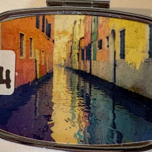 Puede incluir: Un peque&ntilde;o pastillero plateado con tapa abisagrada. La caja presenta una colorida imagen de una escena de canal con edificios en tonos naranja, amarillo y azul. Una peque&ntilde;a pegatina blanca con el n&uacute;mero "14" es visible.