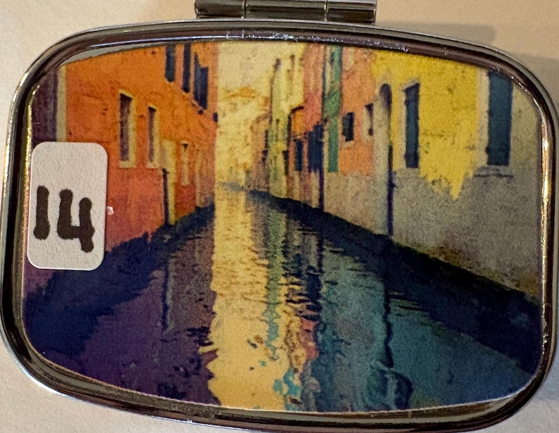 Puede incluir: Un peque&ntilde;o pastillero plateado con tapa abisagrada. La caja presenta una colorida imagen de una escena de canal con edificios en tonos naranja, amarillo y azul. Una peque&ntilde;a pegatina blanca con el n&uacute;mero "14" es visible.