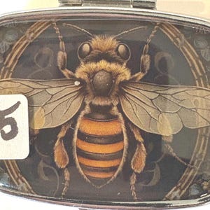 Puede incluir: Un pastillero rectangular plateado con una ilustraci&oacute;n detallada de una abeja. La abeja es marr&oacute;n y naranja, con alas transparentes. El n&uacute;mero "5" est&aacute; en una pegatina blanca en la esquina inferior izquierda. El fondo es oscuro con detalles florales.